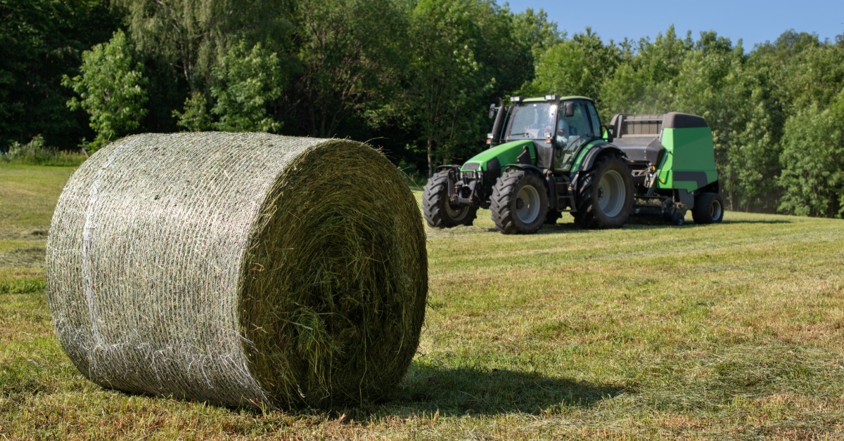 A close-up of a round hay bale in an empty field during the day with a tractor towing a round hay baler behind it.